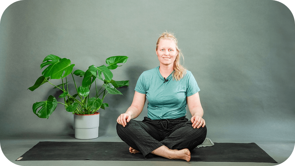 Kursleiterin sitzt lächelnd im Schneidersitz auf einer Yogamatte neben einer Pflanze.
